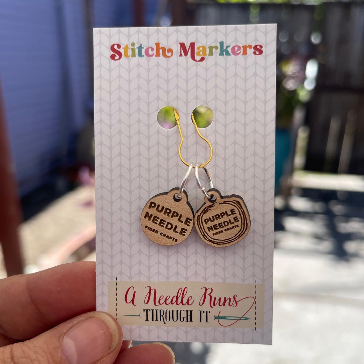 Pair of wooden stitch markers on a card held by a hand with a blurred outdoor background.